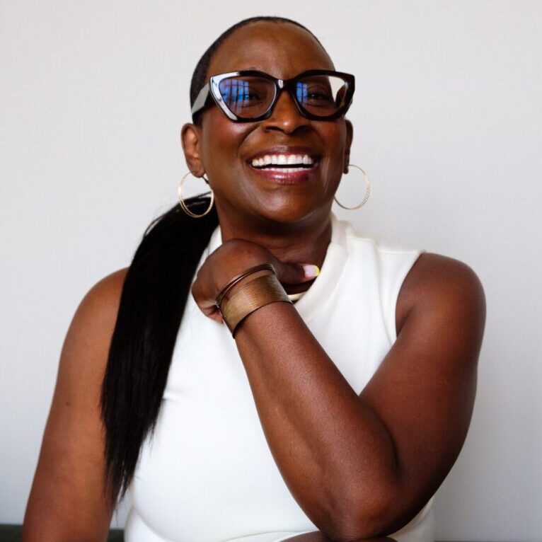 Smiling woman in glasses and white sleeveless top with arm resting on chin.